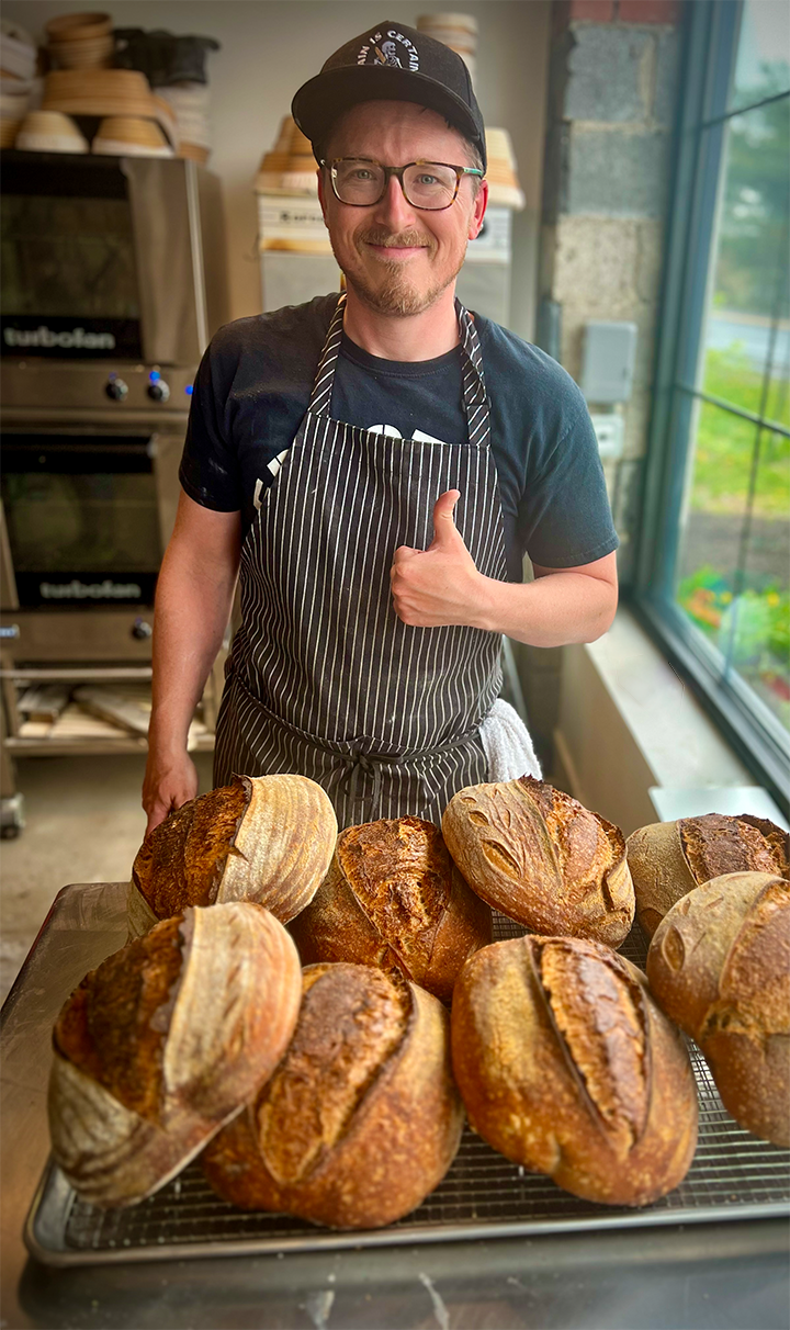 Michael in a striped apron giving a thumbs up behind a tray of fresh sourdough loaves